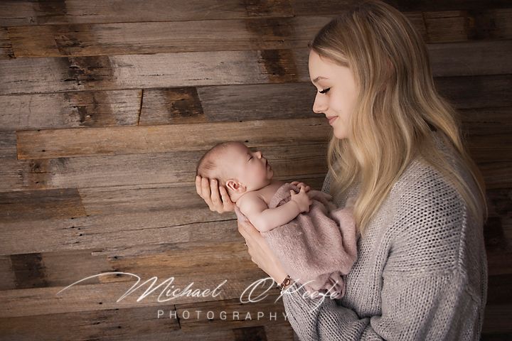A mother tenderly holds her sleeping newborn baby in her hands against a rustic wood plank backdrop.