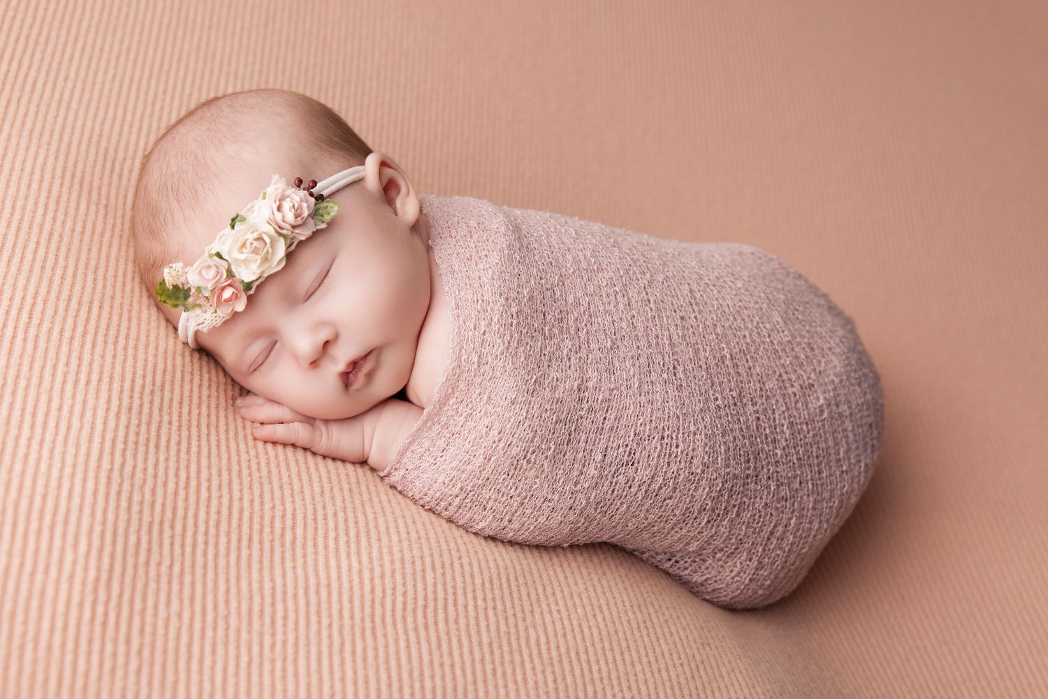 Sleeping newborn wrapped in soft pink swaddle wearing delicate floral headband on peach backdrop.