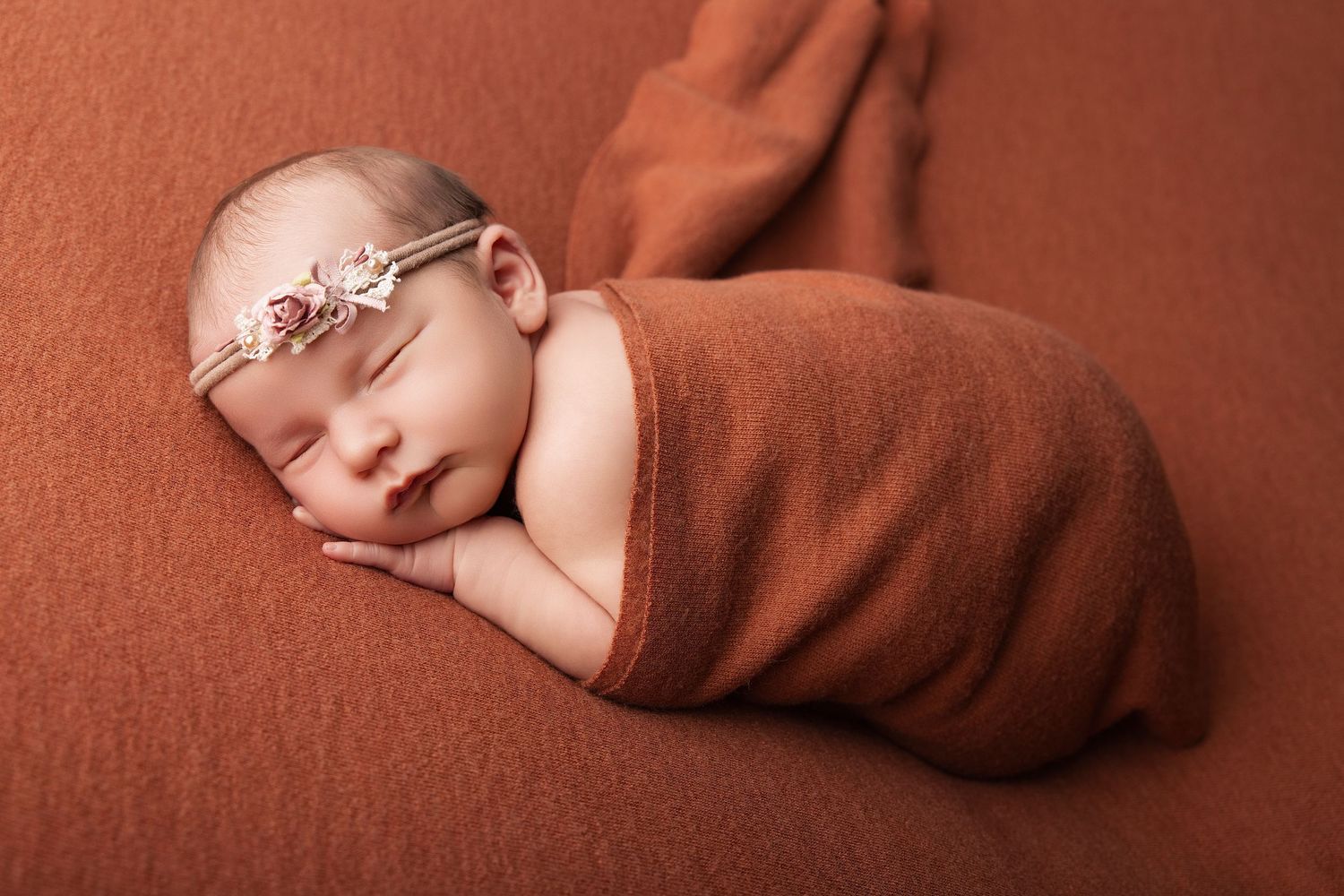Sleeping newborn baby swaddled in rust orange wrap with floral headband on matching terracotta background.