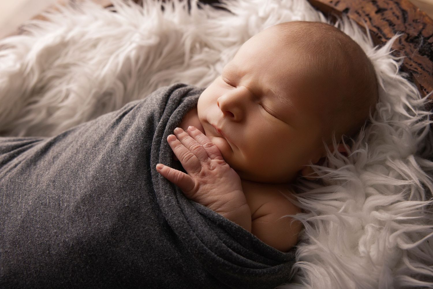 Peaceful newborn baby swaddled in gray wrap sleeping on fluffy white fur, tiny hands visible near face.