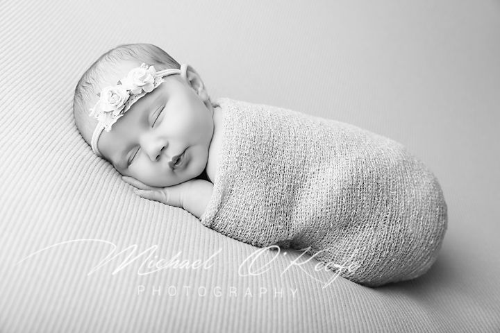 Black and white newborn photo of sleeping baby girl swaddled in soft wrap wearing floral headband.