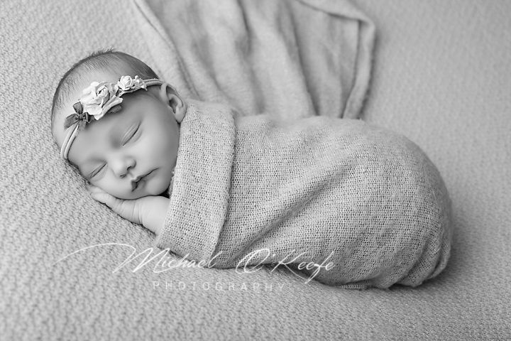 Black and white newborn photo of sleeping baby swaddled in soft wrap wearing floral headband.