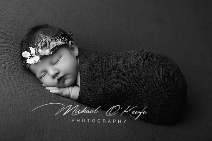 Black and white newborn photo of sleeping baby swaddled in dark wrap with floral headband by Michael O'Keefe Photography.