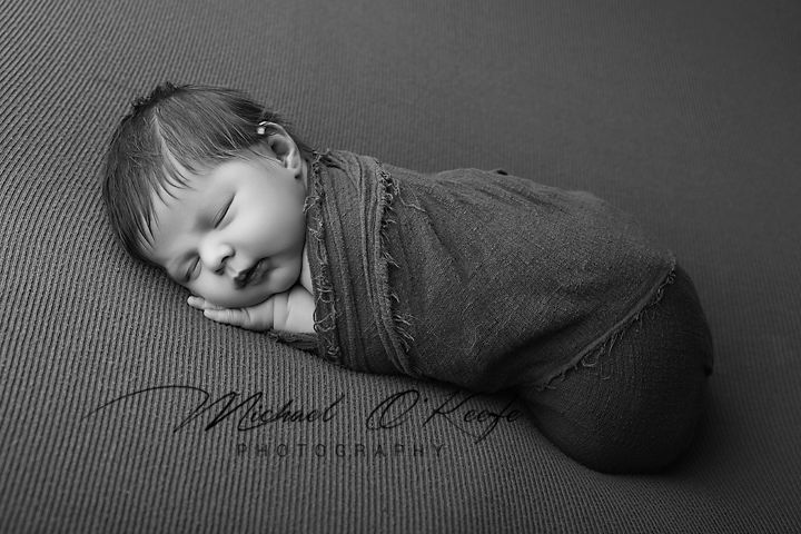 Black and white portrait of a sleeping newborn baby wrapped snugly in a dark blanket on a textured background.
