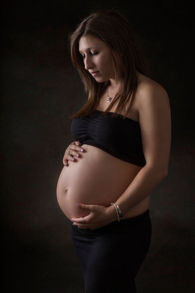 Dramatic studio maternity portrait with woman in black top against dark backdrop.