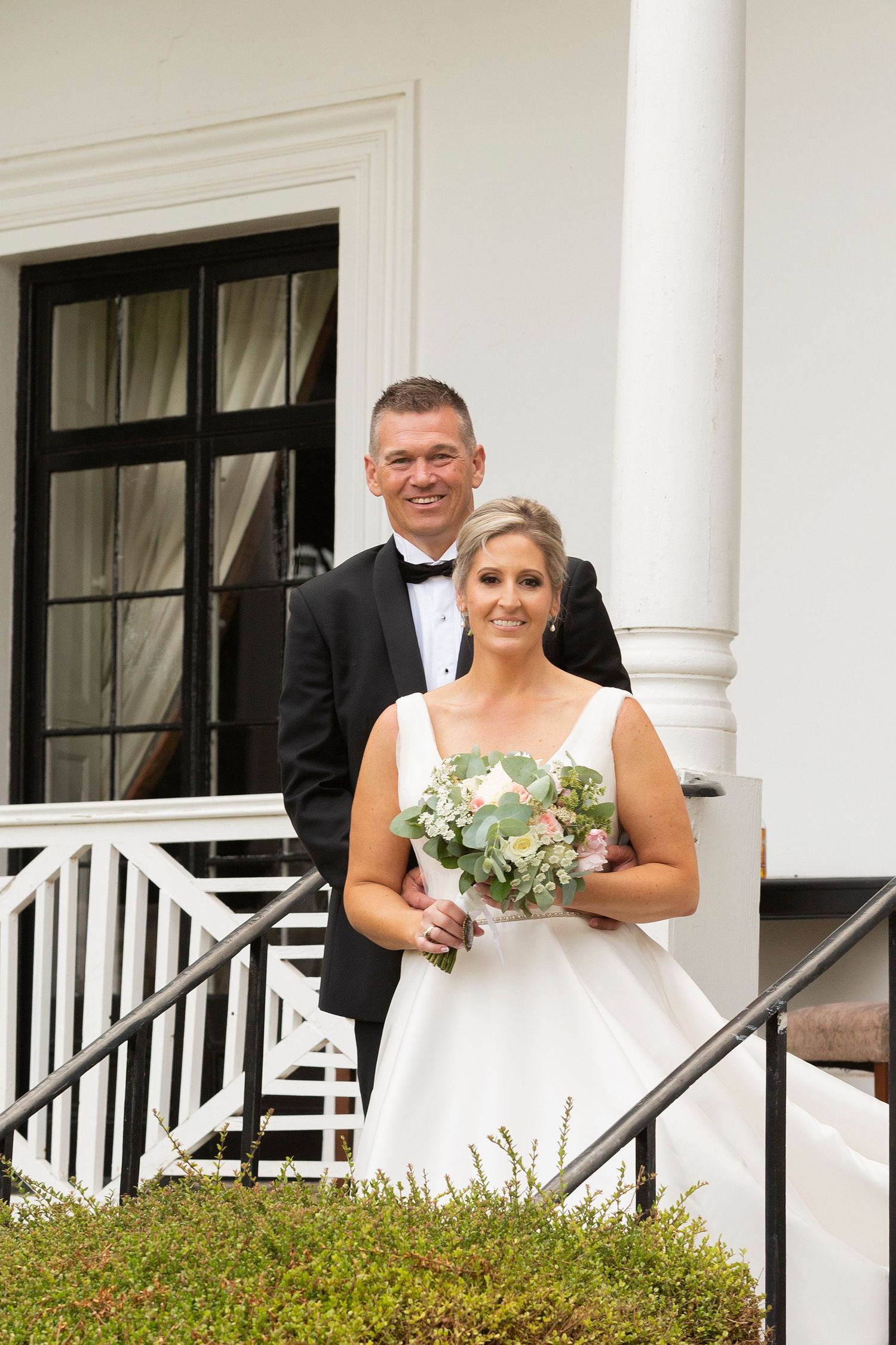 A wedding couple poses on white porch steps of a historic building with elegant columns.