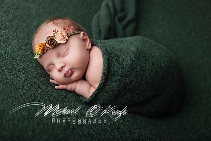 Newborn baby girl swaddled in green wrap with floral headband, sleeping peacefully in professional portrait photography.