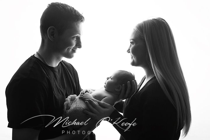 Black and white newborn portrait of smiling parents facing each other while gently holding their sleeping baby in silhouette.