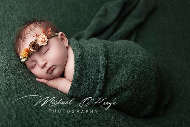 Sleeping newborn baby girl wearing a floral headband, wrapped in a green knit blanket on a green backdrop in a newborn photography portrait.