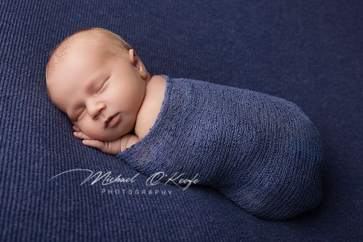 Newborn baby swaddled in a blue knit wrap, sleeping peacefully on a matching navy blue textured background.