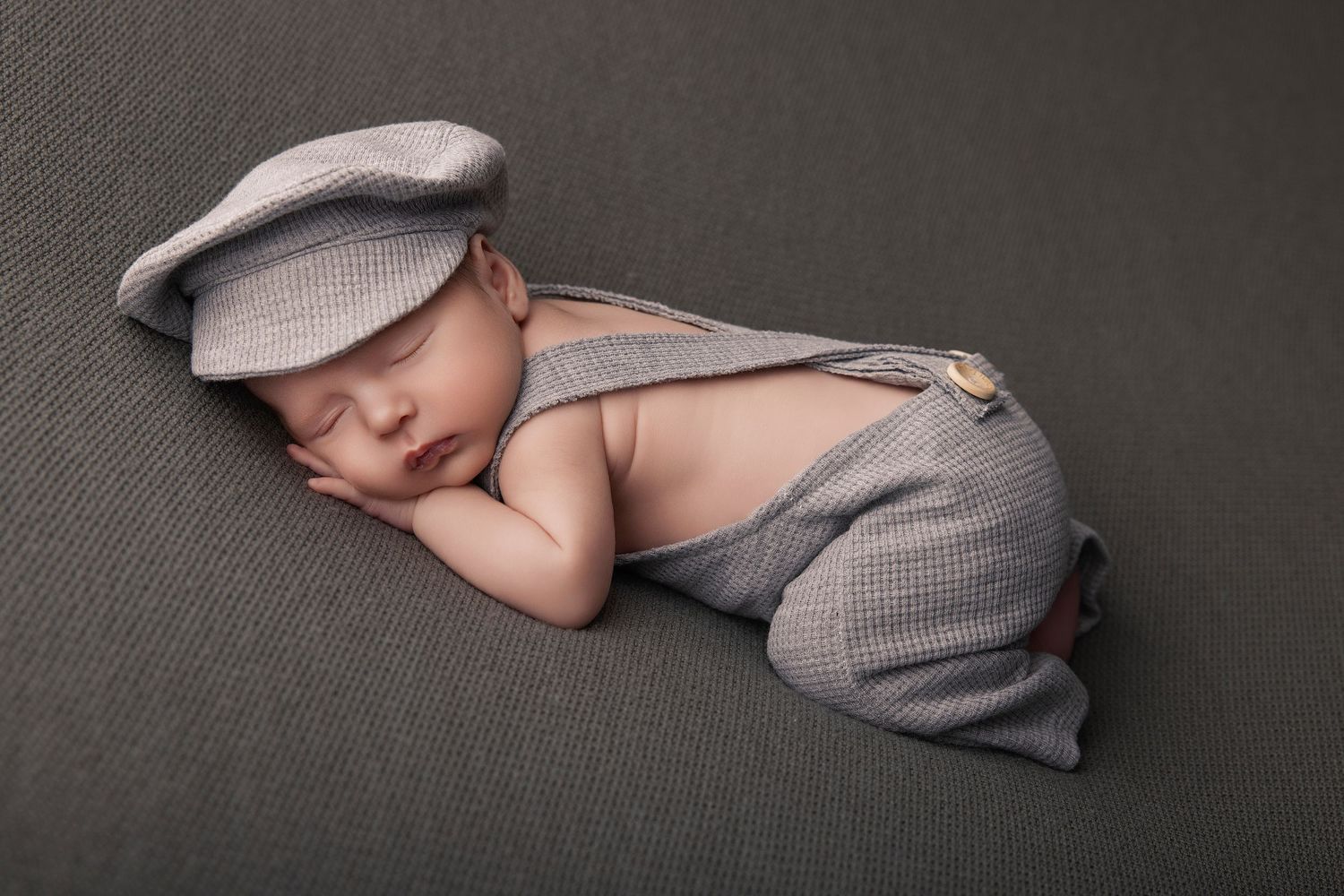 Newborn baby sleeping peacefully in gray suspender pants and matching flat cap on a gray background.