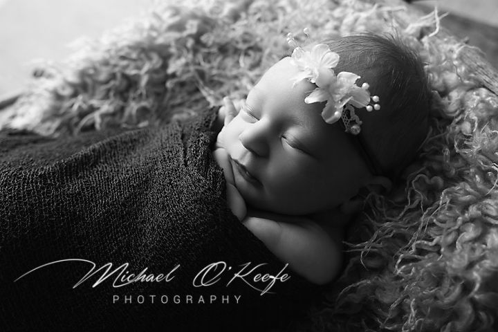 Black and white newborn photo of sleeping baby wrapped in blanket with floral headband by Michael O'Keefe Photography.