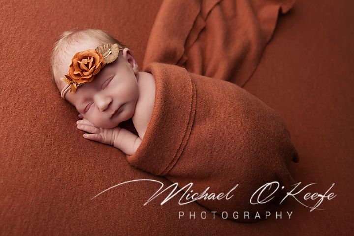 Newborn baby girl sleeping peacefully wrapped in rust orange knit blanket with floral headband by Michael O'Keefe Photography.