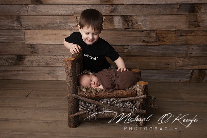 Older toddler boy in black shirt sits beside swaddled newborn baby in rustic wooden bed, professional studio photo.