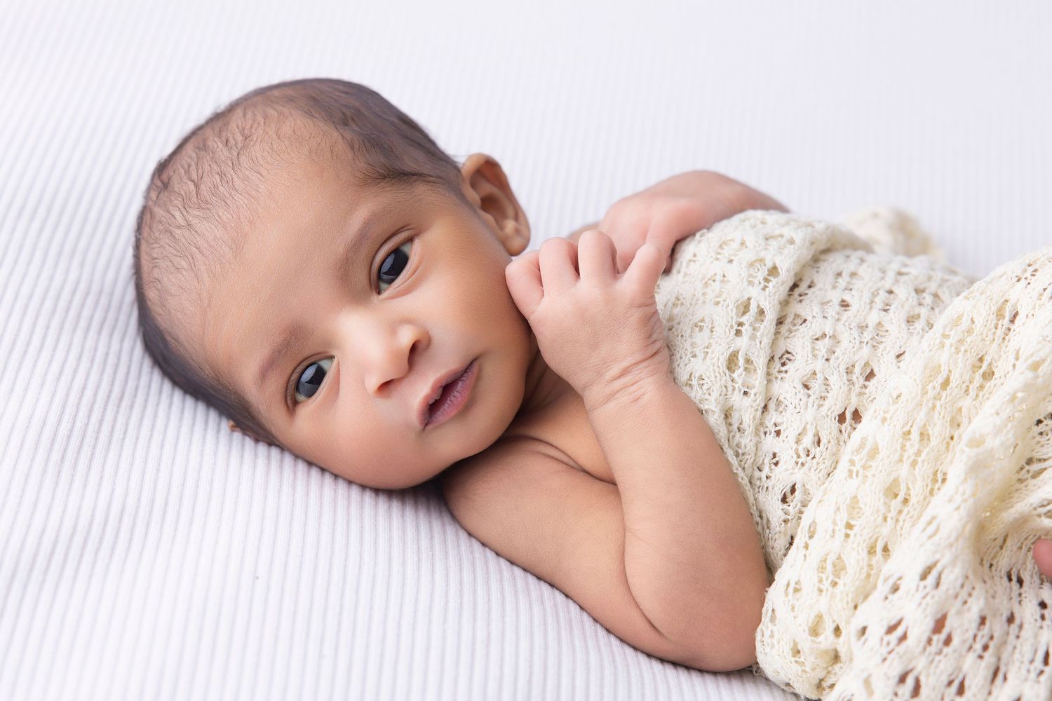 Small infant partially covered with cream knit blanket on white textured background.