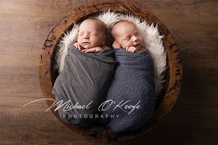 Two swaddled newborns sleep peacefully together in a wooden bowl lined with soft white fur.