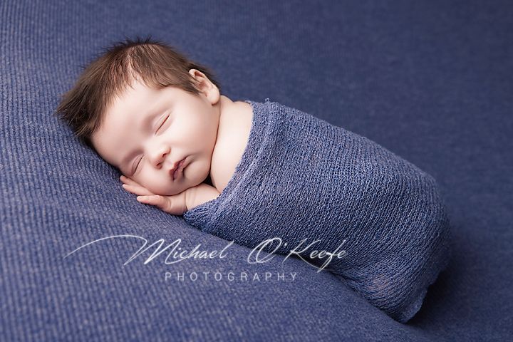 A sleeping newborn swaddled in a gray wrap rests peacefully on dark blue fabric during a professional photo session.