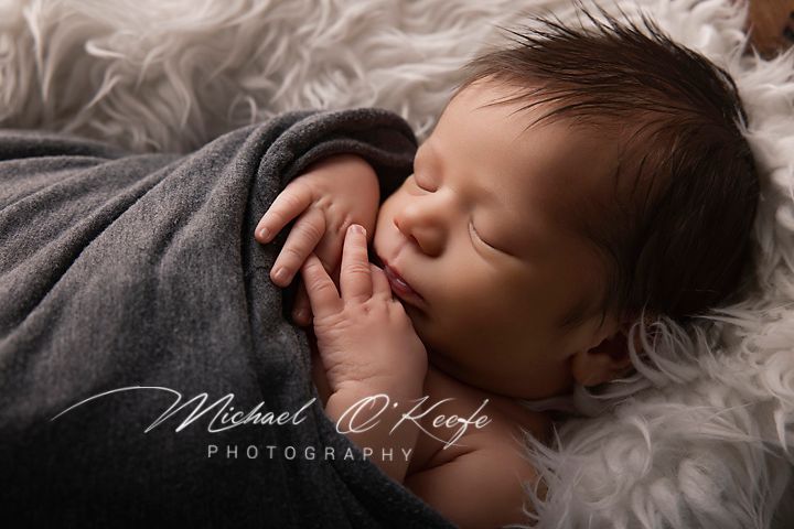 Sleeping newborn baby wrapped in gray swaddle on white fluffy blanket, professional newborn photography by Michael O'Keefe.
