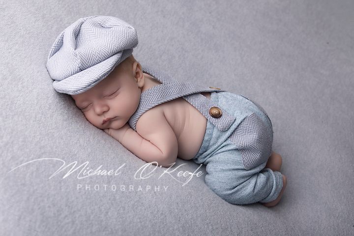 Sleeping newborn baby wearing a light blue knit cap and suspender outfit on a gray background.