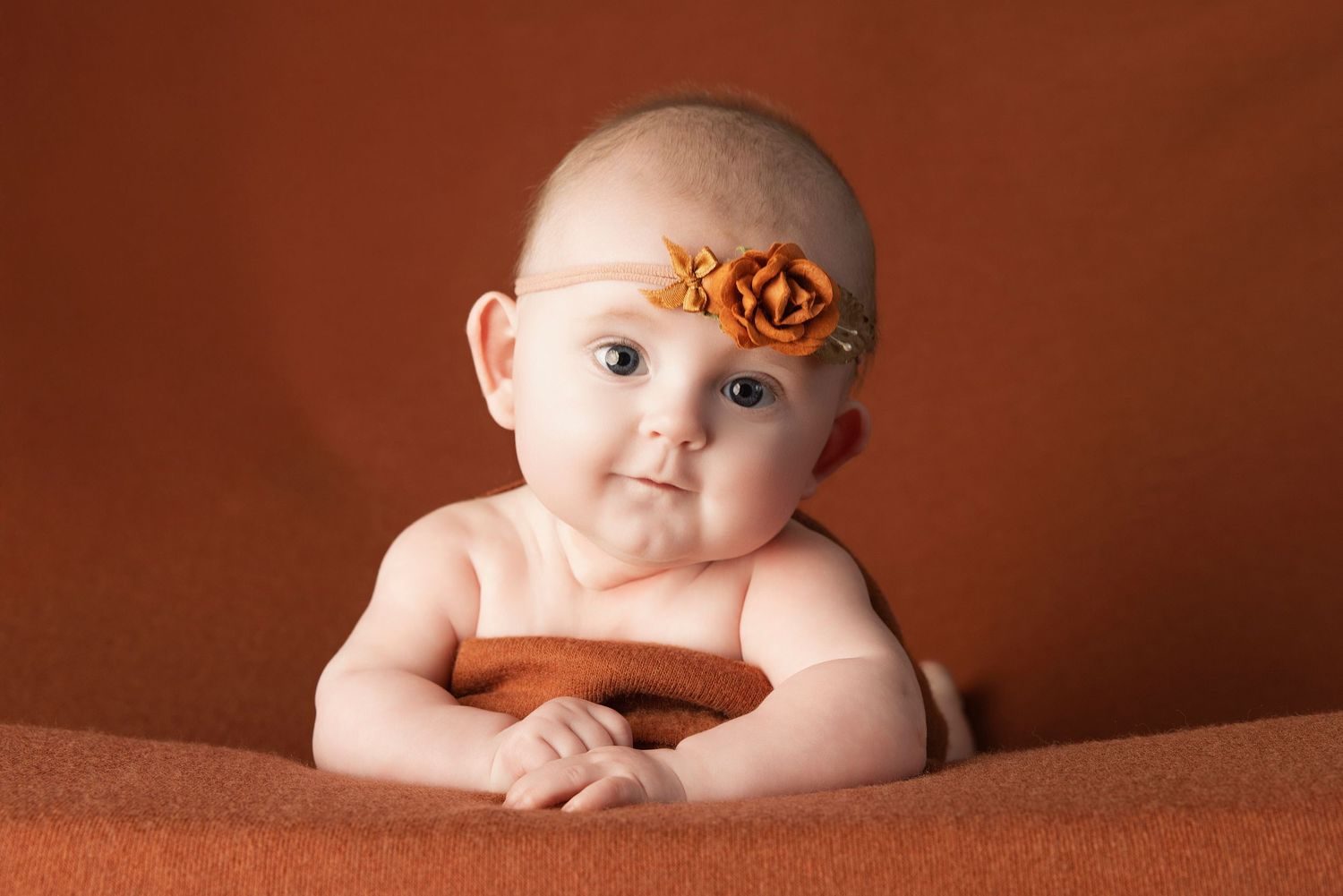 A baby lies on a brown blanket with orange flower hair accessories in a series of artistic portrait poses.
