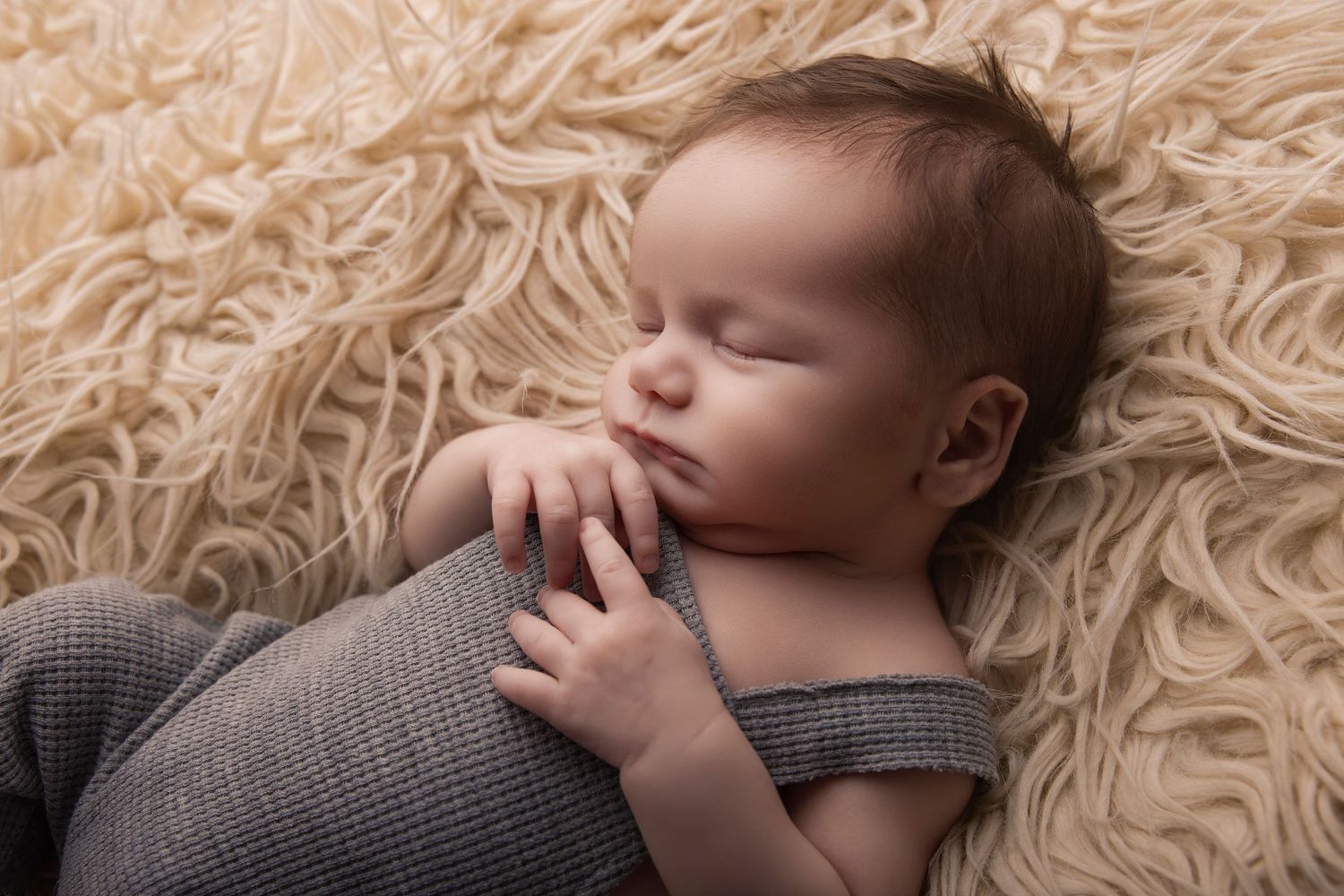 Sleeping newborn baby in gray knit overalls resting peacefully on a soft cream fluffy blanket.
