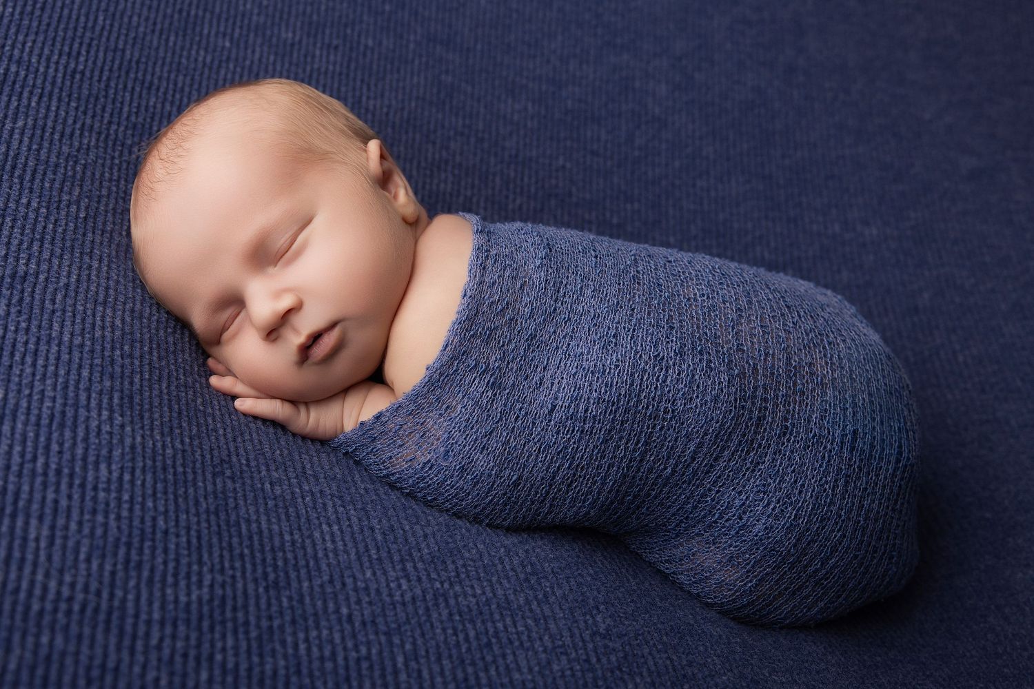 Newborn baby swaddled in a blue knit wrap, sleeping peacefully on a navy blue textured background.
