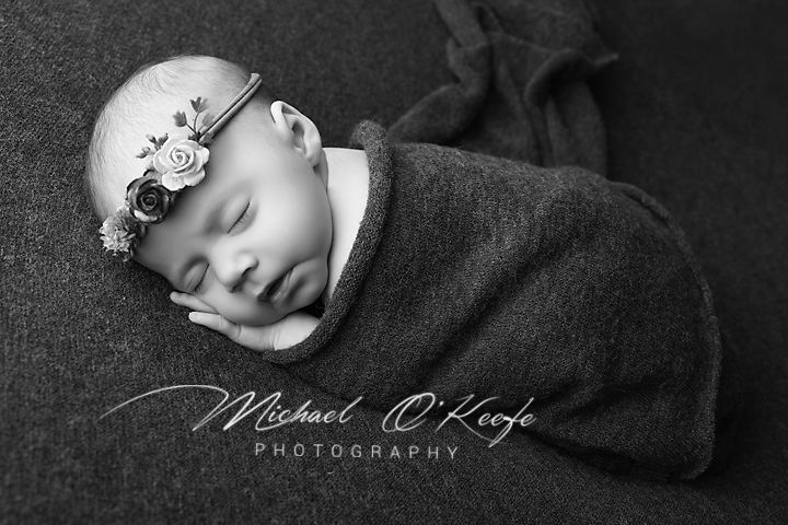 Black and white newborn photography of sleeping baby wrapped in gray swaddle with floral headband by Michael O'Keefe.