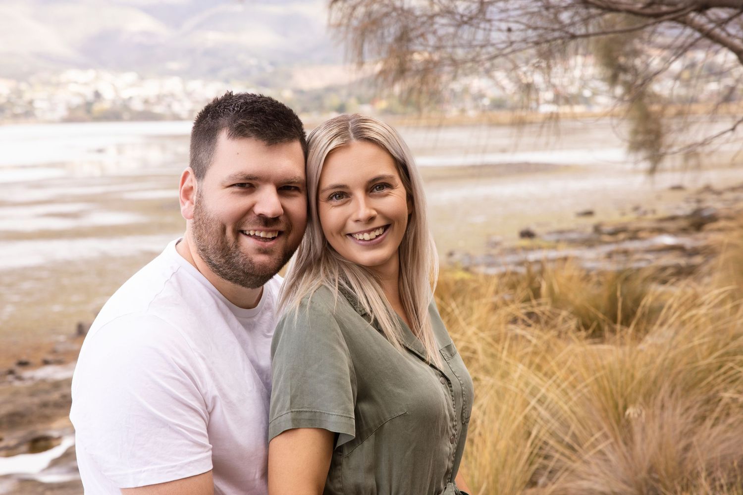 Couple smiling together outdoors with waterfront and natural landscape in background.