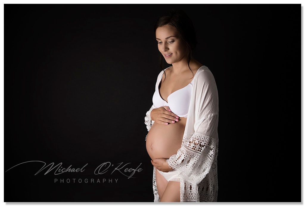 Artistic black and white maternity portrait with woman in white robe against dark background.