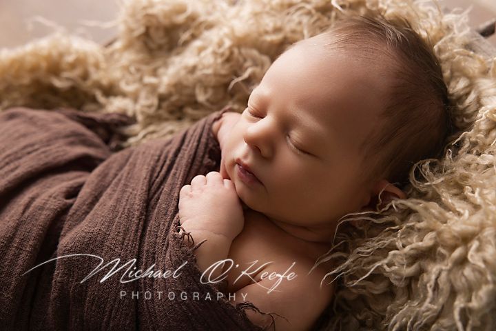 Newborn baby sleeping peacefully wrapped in brown cloth on fluffy tan fur, professional infant photography by Michael O'Keefe.
