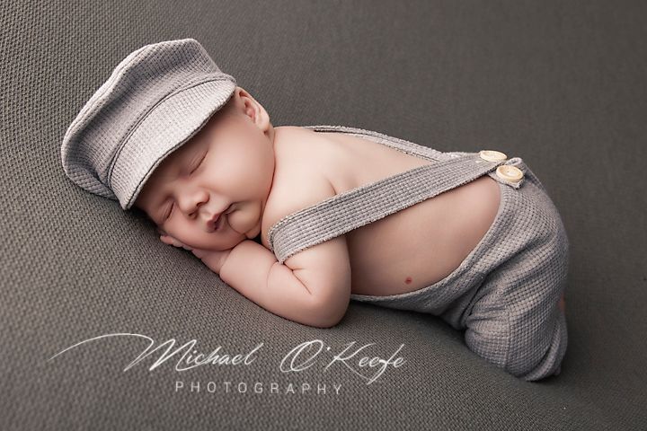 Newborn baby sleeping peacefully in gray suspender outfit and matching newsboy cap on gray backdrop.
