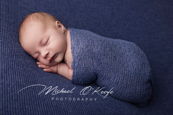 Newborn baby sleeping peacefully wrapped in a blue knit blanket, professional photography by Michael O'Keefe.