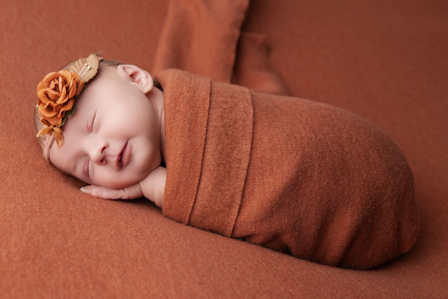 Sleeping newborn baby wrapped in brown fabric with a matching flower headband.
