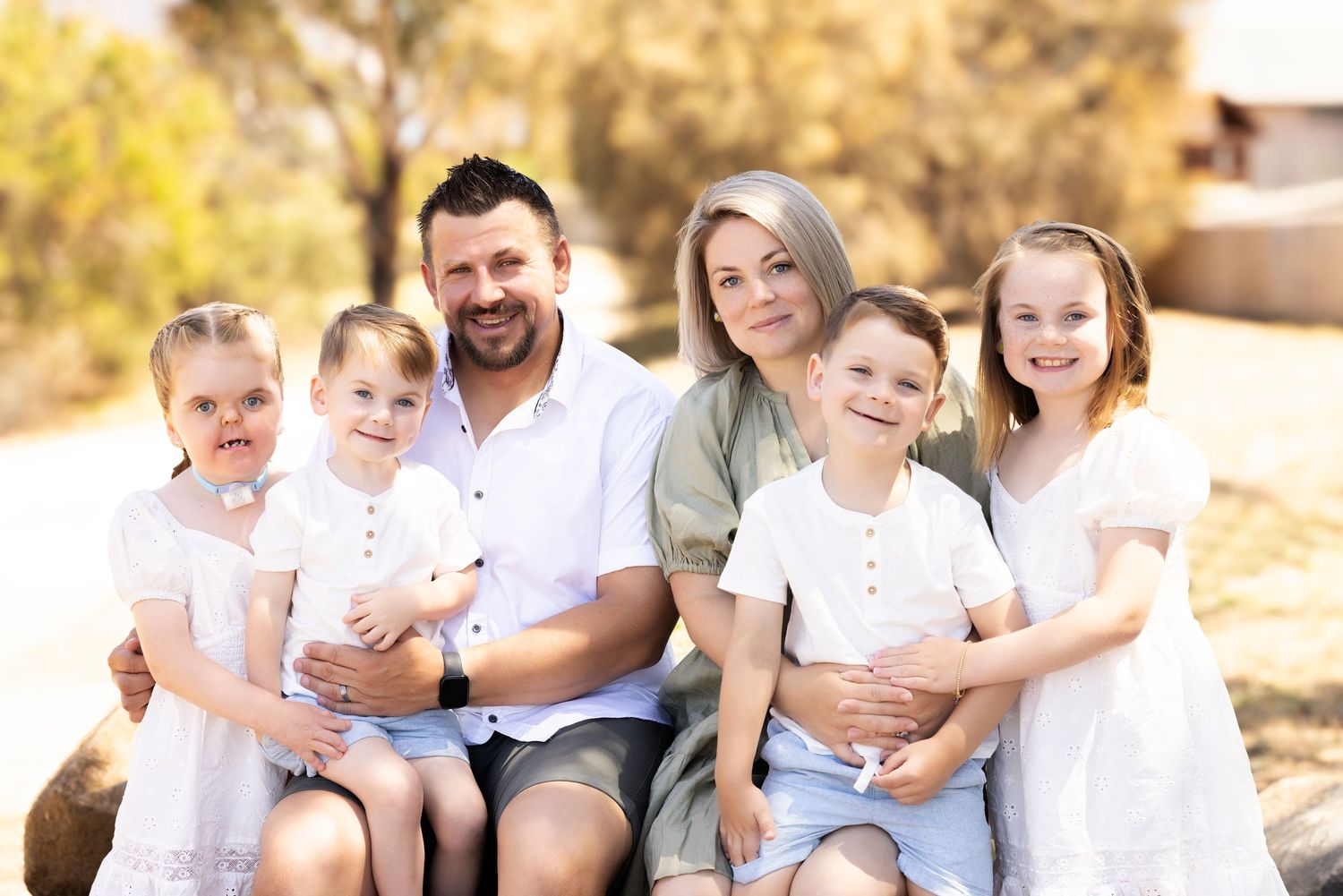 Family portrait of five people sitting together outdoors in coordinated white outfits.