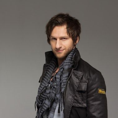 Person in black jacket and scarf poses dramatically against gray backdrop.
