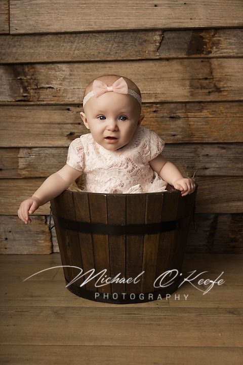 Adorable baby girl in pink lace outfit sitting in rustic wooden barrel bucket, professional studio portrait photography.