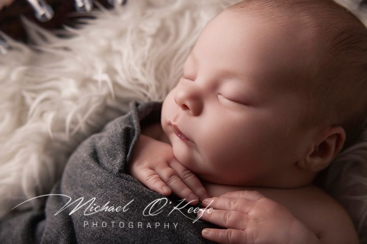 Newborn baby sleeping peacefully on fluffy white fur, wrapped in gray fabric, in a professional photography studio portrait.