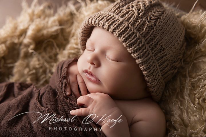 Peaceful newborn baby sleeping in a knit brown hat, nestled in soft fur, captured by Michael O'Keefe Photography.