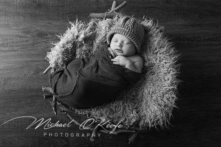 Black and white newborn photo of sleeping baby wrapped in dark fabric, wearing knit hat, nestled in rustic basket with fur.