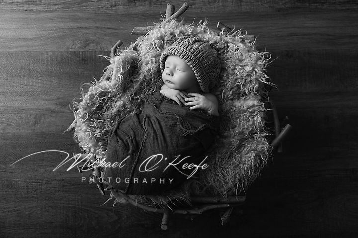 Black and white newborn photography of sleeping baby in knit hat nestled in rustic wooden bowl by Michael O'Keefe.