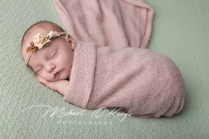 Newborn baby swaddled in pink wrap sleeping peacefully on mint green backdrop with floral headband.
