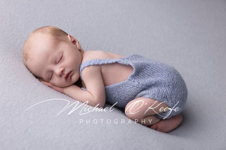 A sleeping newborn baby in soft grey wrapping lies peacefully against a light backdrop during a professional photo session.