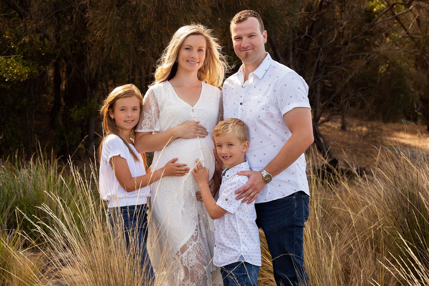 Family of four poses outdoors in golden light, pregnant mother in white dress with husband and two young children.