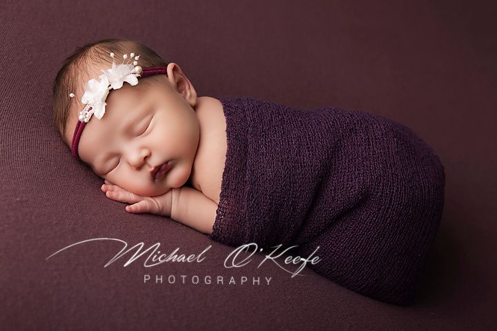 Newborn baby swaddled in purple wrap with floral headband, sleeping peacefully on purple backdrop.
