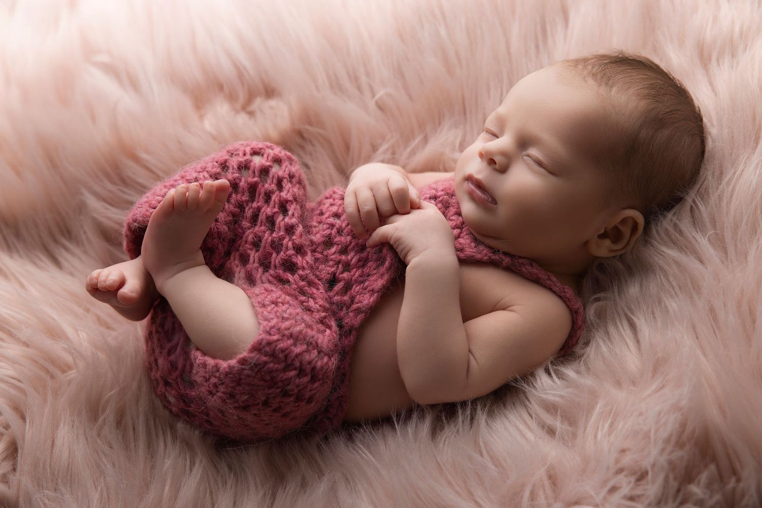Newborn baby wearing pink lace romper resting on soft pink fur blanket in artistic studio photography poses.