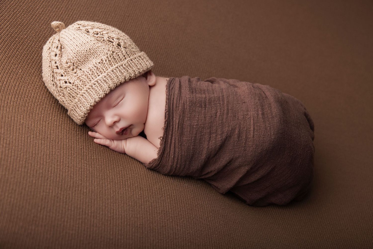 Newborn baby sleeping peacefully swaddled in brown wrap wearing a knit beige hat on brown background.