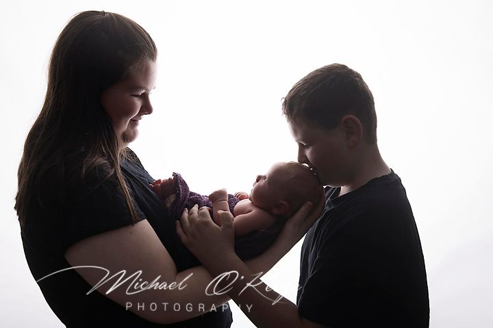 A mother and older sibling lovingly hold a newborn baby in a beautiful backlit portrait by Michael O'K Photography.