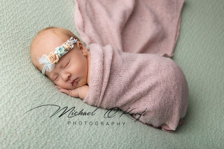 Newborn baby girl swaddled in pink wrap with floral headband sleeping on mint green backdrop.