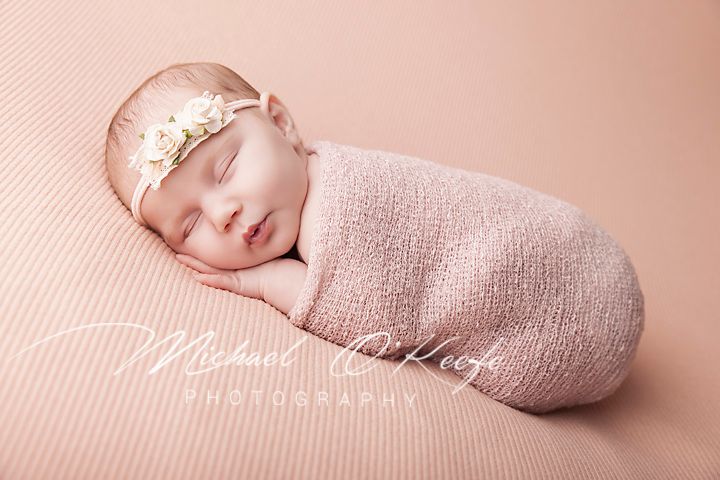 Sleeping newborn baby girl swaddled in a soft pink wrap with a floral headband on a pink striped background.