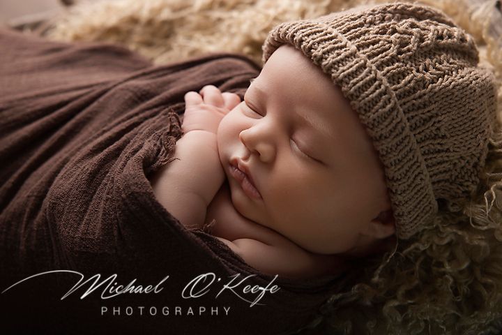 A sleeping newborn baby wrapped in brown fabric with a knit hat lies peacefully during a professional photo session.